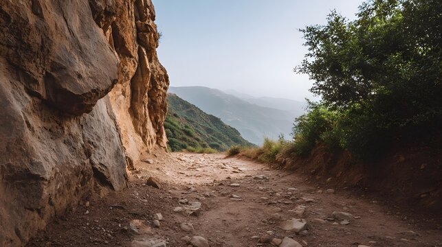 A dusty rocky mountain trail ascends past a towering cliff face toward distant hazy peaks and a clear sky