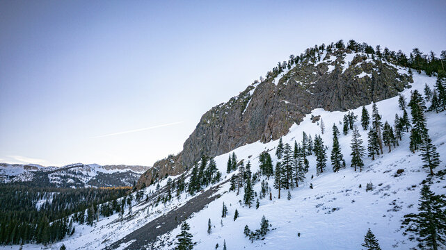 Aerial view of snow-dusted mountainside, where stark rock faces emerge amidst a tapestry of evergreen trees, Mammoth Lakes, California, United States.