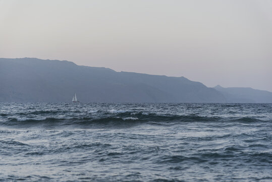 Lonely sailboat sailing on rough sea against misty mountains at twilight