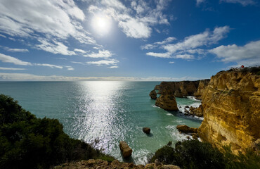 A dramatic sunset seascape with dark rocky cliffs. Golden sunlight breaks through dramatic storm clouds, creating bright orange and yellow highlights again