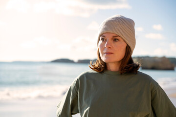 Woman standing on sunny beach wearing a knit hat looking towards the horizon