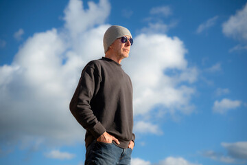 stylish man wearing knit hat and sunglasses stands with hands in pockets, looking into the distance