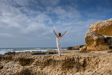 woman standing on rocks by the ocean with arms raised meditating and practicing yoga