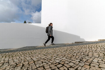 Woman walking along white wall on cobblestone street with backpack