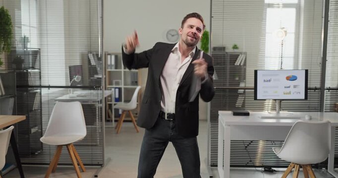 Happy businessman manager dancing in office celebration. He celebrates results in a modern workspace with glass walls, smiling near charts on the screen. Concept shows corporate success and joy.
