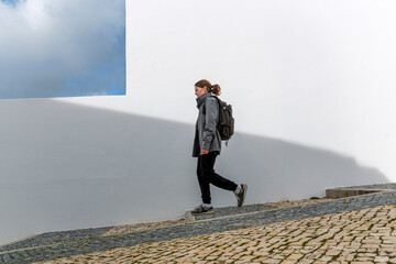 Woman walking along white wall on cobblestone street with backpack