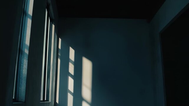 Sunlight beams stream through a window, casting strong geometric shadows and bright light patterns on a minimalist wall and dark floor in an empty room, creating a sense of quiet and contemplation
