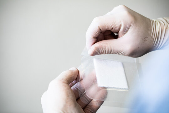 A healthcare worker peels the wrapping off of a hydrocolloid bandage against a white wall in a brightly lit healthcare setting hospital 