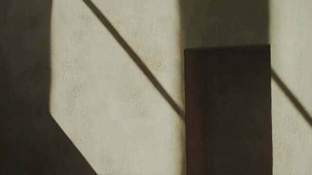 Beige wall featuring a recessed niche, illuminated by warm sunlight creating sharp, diagonal patterns of light and shadow, emphasizing minimalist architecture and textured surfaces