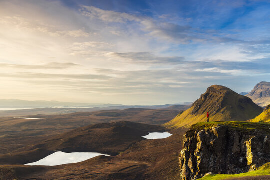 Aerial view of a lone figure perched on a rugged cliff edge overlooking the vast, textured landscape under a vibrant sky, Portree, Scotland, United Kingdom.