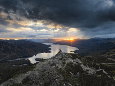 Aerial view of the rugged peak overlooks a tranquil loch, as the setting sun paints the sky in hues of gold and umber, Callander, Scotland, United Kingdom.