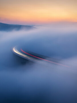 Aerial view of a bridge piercing through a dense fog blanket, light trails painting streaks across the misty canvas, Crni Kal, Koper, Slovenia.