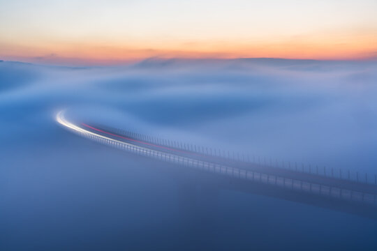 Aerial view of a bridge disappearing into a sea of fog under a pastel sky, the road's lights streak through the dense whiteness, Crni Kal, Koper, Slovenia.