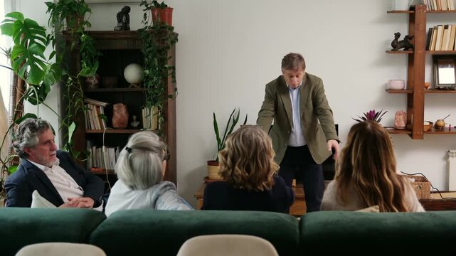 Middle aged man energetically gestures while friends on a sofa guess during a game of charades