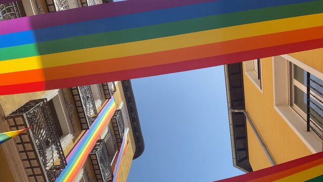 Colorful rainbow flags hanging across a sunny street, between yellow building facades, celebrating diversity, equality, and pride in madrid