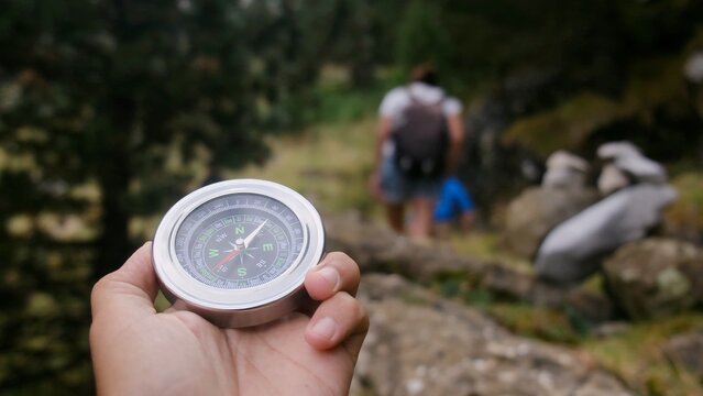Hand holding a bright silver compass pointing the way forward while a hiker with a backpack walks blurred in the background, venturing into a natural landscape