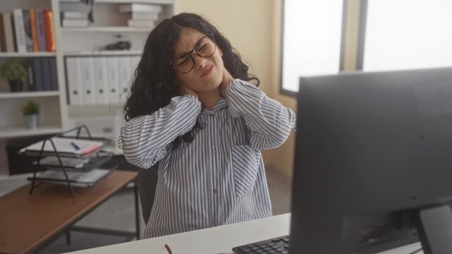 Young woman with glasses holds neck with both hands at office desk beside a computer monitor, wincing from neck pain; work stress.
