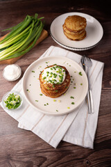 stack of golden potato pancakes topped with sour cream and chopped green onions on a ceramic plate, rustic wooden table setting with extra pancakes and cutlery, cozy breakfast scene with warm lighting