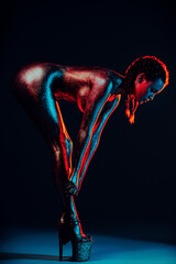 Woman bending toward her legs on black studio background with blue, red, and pink lighting, wearing high heels, paint on body