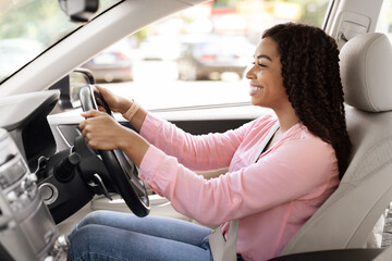 Side view profile portrait of joyful African American woman driving car, doing test drive in dealership showroom. Happy lady riding brand new nice car, sitting on driver's seat in luxury automobile © Prostock-studio
