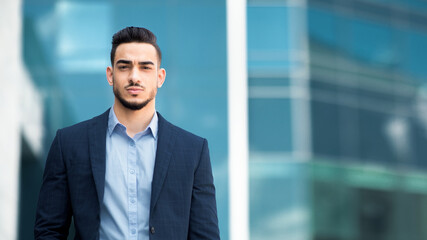 Fototapeta premium Handsome middle-eastern young businessman in suit posing next to office center, serious arabic entrepreneur having break after successful busienss meeting, standing on street, copy space, panorama
