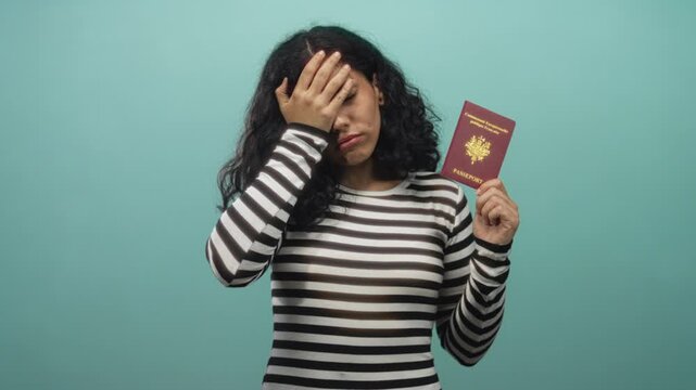 Woman holding passport and covering face with hand to forehead in studio with teal backdrop; travel frustration.