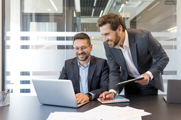 Two smiling business professionals working together on a laptop during an office meeting, discussing projects, analyzing data, and finding solutions for successful teamwork and strategic planning