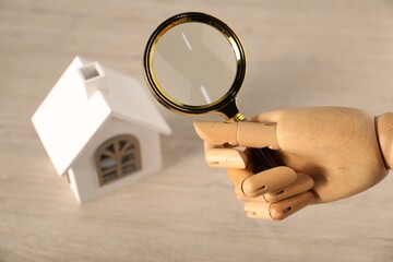 Mannequin hand with magnifying glass near house model at wooden table, above view