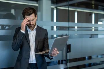 Fotobehang Onderzeeër Stressed businessman in a suit suffering from a headache and work pressure while holding a laptop, standing by a frosted glass partition in a modern office environment  © Liubomir