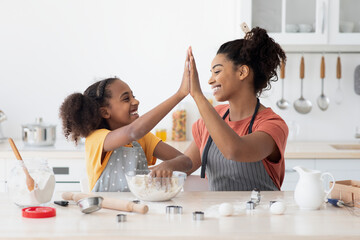 Cheerful black mother and daughter giving high five while cooking at home, wearing aprons, happy...