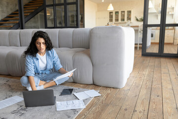 Focused young woman working online, studying financial documents, using laptop computer at home,...