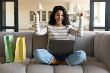 Excited young woman with laptop and shopping bags making YES gesture, buying things online from...