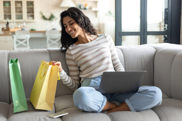 Cheery young woman sitting on couch with laptop, looking inside shopper bags, ordering goods online...