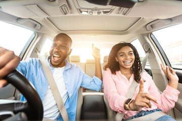 Road Trip Concept. Front view portrait of excited playful African American couple driving luxury...