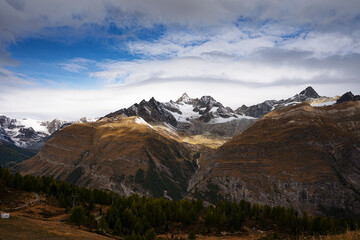 Fototapeta premium Panoramic view of snow-capped mountain peaks in the Alps with dramatic sunlight hitting the brown slopes under a cloudy blue sky with pine forests.