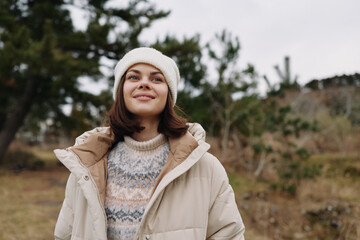 Woman outdoors in a beige coat and knit sweater, smiling in a soft winter mood against a natural...