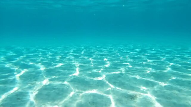 Sunlight filters through clear turquoise water onto a sandy seabed. Gentle waves create shimmering patterns on the ocean floor.