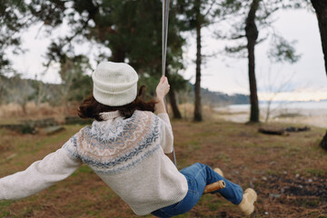Swinging outdoors among trees by a lake, a person in a sweater and beanie enjoys a peaceful moment...