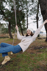 Girl on a swing in an outdoor park, smiling and joyful, wearing a beanie and sweater, capturing carefree youth and playful mood in a natural setting
