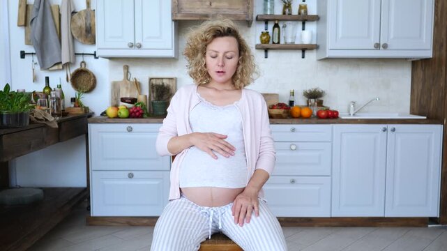 Young pregnant woman experiencing labor contractions, breathing deeply while sitting in her kitchen at home. Mother to be is preparing for childbirth and practicing relaxation techniques