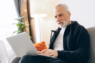 Portrait of smiling senior man sitting on comfortable sofa at home using laptop, checking mail