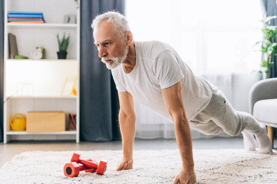 Senior bearded gray haired man doing exercise standing in plank, push up at home in cozy living room