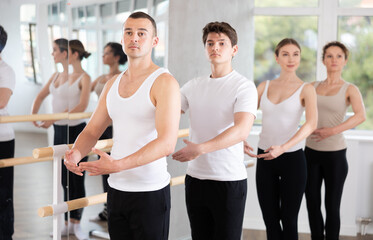 Fototapeta premium Sports young man in black and white sportswear, training first position of ballet stance at barre with group in bright choreography studio