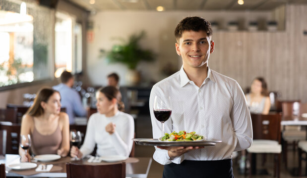 Young male waiter posing with tray of salad and wine in restaurant