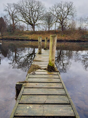 Fototapeta premium Old wooden jetty extending into a calm river with mirror-like reflections of bare winter trees.