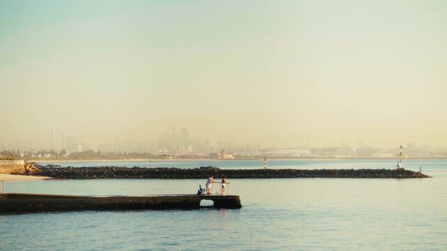 People Relaxing on Ramsgate Beach Pier with Calm Sea and Sydney City Skyline in Hazy Sunrise Background