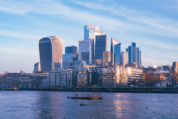 Morning light bathes the modern skyline of London as the Thames river gently flows by. Skyscrapers rise above the city, creating a stunning urban landscape.