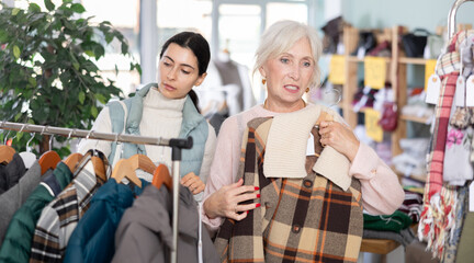 Obraz premium Two women in the store are looking at a warm coat and down jackets, discussing the quality and style among the assortment of outerwear. Mature woman and young woman buying warm clothes in a mall
