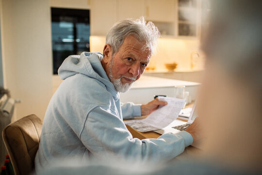 Senior man paying bills on laptop at home kitchen