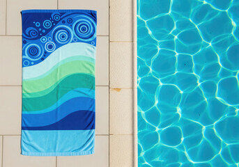 Obraz premium Top view of a blue and green patterned beach towel on a tiled poolside next to crystal clear swimming pool water.